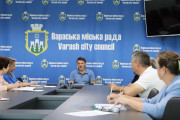 Засідання Інвестиціїної ради у Вараській міській раді. На задньому плані синій банер з гербом міста та написом "Вараська міська рада Varash city council". За великим столом сидять кілька людей, обговорюючи щось.