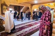 Священик стоїть обличчям до групи людей у дерев'яній церкві та читає молитву. На задньому фоні група людей, котрі стоять, схиливши голови донизу. Жінка, що стоїть найближче до камери, у хустці, одягнена у сукню з квітковим візерунком. Навколо них видно дерев'яні стіни та ікони.