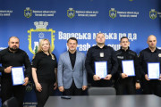 На фотографії зображено групу людей, серед яких працівники поліції та посадовець, що стоять на фоні синього банера з написом «Вараська міська рада / Varash city council». Четверо осіб у поліцейській формі тримають у руках грамоти за досягнення. У центрі стоїть чоловік у сірому піджаку, який є представником міської влади. Зліва — жінка у чорній формі з емблемою, можливо, також співробітниця поліції. Атмосфера офіційна, церемонія нагородження.