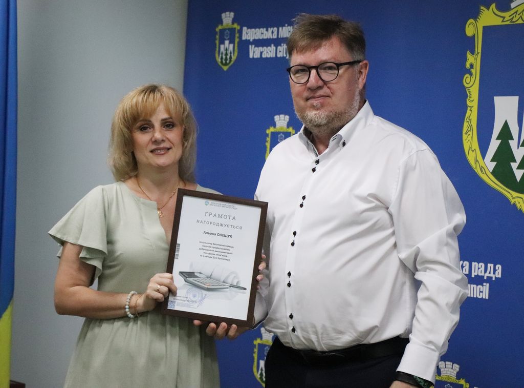 На фото Олександр Мензул нагороджує працівника бухгалтерії освітнього закладу. Позаду банер Вараської міської ради.