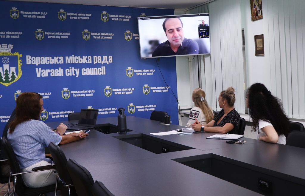 Група людей сидить за великим столом. На задньому плані синій банер з написом "Вараська міська рада" та гербом міста. На екрані телевізора, закріпленого на стіні, видно чоловіка, який бере участь у зустрічі онлайн.