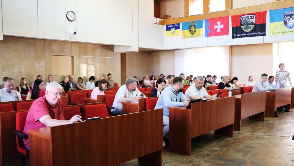 На цьому зображенні показано велике приміщення, де проходить засідання 50 сесії. У залі розташовані ряди дерев'яних столів з червоними кріслами, які займають депутати ВМР. Деякі з них дивляться вперед, інші зосереджені на своїх записах або пристроях. На задньому плані, над сценою або президією, висять прапори, серед яких можна розрізнити прапор України та інші стяги. 