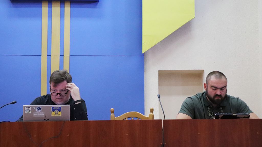 На цьому знімку крупним планом зображено міського голову м.Вараш Олександра Мензула та секретаря ВМР - Геннадія Дерев'янчука за столом. За ними видно синю стіну з жовтими вертикальними смугами та елементами інтер'єру.