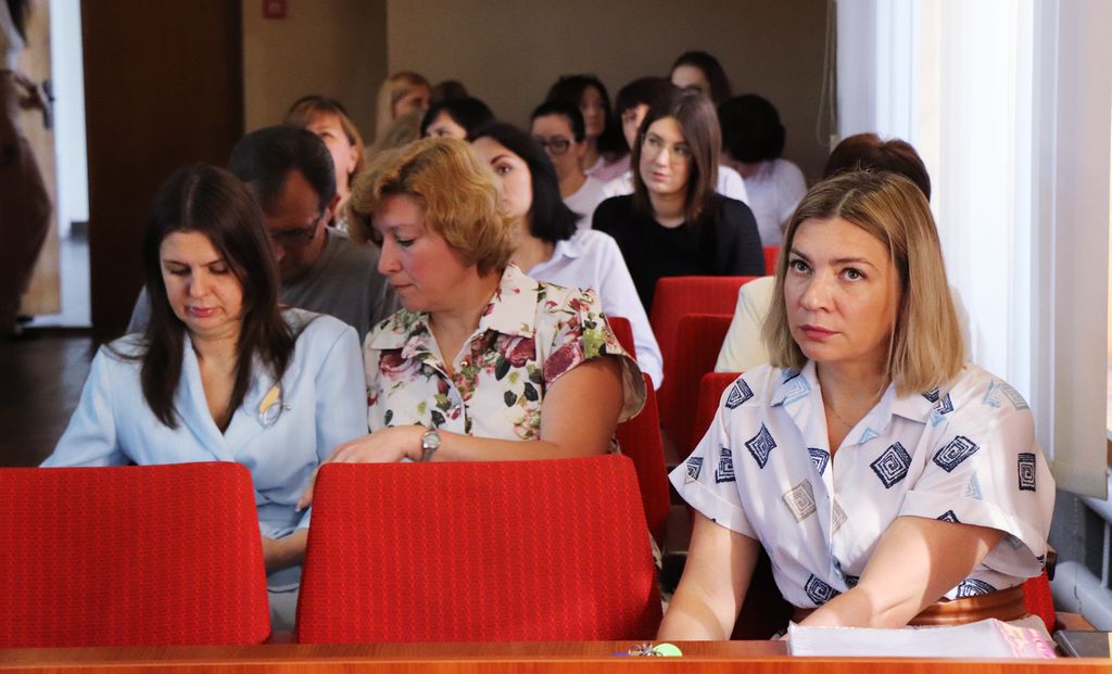 Ця фотографія є крупним планом працівників виконкому, що сидять у червоних кріслах на засіданні 50 сесії. На передньому плані видно трьох жінок, які зосереджено дивляться вперед або вбік. Жінка праворуч одягнена у світлу сорочку з візерунком і має світле волосся. Жінка в центрі також у світлій блузці з квітковим принтом. Жінка ліворуч у світлій блузці. За ними видніються інші учасники засідання, обличчя яких менш чіткі.