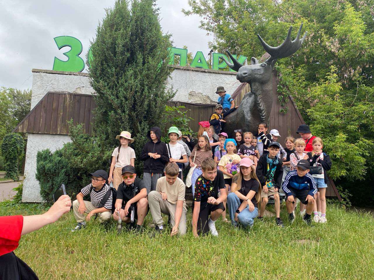 Група дітей та кілька дорослих позують для фотографії перед будівлею. Праворуч від будівлі розташована велика темна статуя лося. Група складається переважно з дітей шкільного віку, одягнених у повсякденний літній одяг, деякі в головних уборах. Вони стоять та сидять навпочіпки на трав'янистій ділянці. На задньому плані видно дерева та захмарене небо.