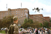 На фотографії зображено чоловіка у військовій формі, який стоїть спиною до глядача та дивиться на дрон, що летить у повітрі. Чоловік одягнений у камуфляжну куртку та кепку.  Дрон чорного кольору з чотирма пропелерами та камерою знизу. Він завис у повітрі над групою людей, які зібралися на вулиці на ярмарок. Видно торгові прилавки, а також припарковані автомобілі вздовж дороги.  На задньому плані розташовані кілька багатоповерхових будинків бежевого та червоного кольорів. 