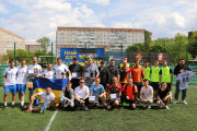 На фотографії зображена велика група чоловіків на футбольному полі зі штучним зеленим покриттям. Вони позують для групового знімка у два ряди, тримаючи футбольний м'яч, український прапор та нагородні атрибути.  У другому ряду стоять чоловіки, багато з яких мають на шиї медалі різного ґатунку (золоті, срібні, бронзові) та тримають дипломи. Один чоловік тримає розгорнутий український прапор.  У центрі другого ряду знаходиться заступник міського голови Дмитро Стецюк. Він одягнений у сіру футболку поло та тримає срібну нагородну табличку.  На задньому плані видно огорожу футбольного поля, багатоповерхові будинки та великий синьо-жовтий банер з написом "ГРАЙ ДЛЯ ЗСУ" та зображенням футболістів. Небо на фотографії світле, з невеликими хмарами.  Загальна атмосфера на фотографії святкова та піднесена, що відповідає події нагородження переможців спортивного турніру.