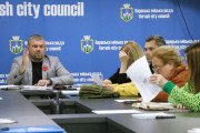 На фотографії зображено засідання комісії у Вараській громаді. За довгим столом, вкритим сірою поверхнею, сидять кілька людей. У центрі ліворуч сидить Дмитро Ющук, директор Департаменту житлово-комунального господарства, майна та будівництва, одягнений у сірий піджак в клітинку, чорну футболку та з червоною квіткою-маком на лацкані. Він жестикулює правою рукою, щось пояснюючи. Перед ним на столі лежать документи та, можливо, мікрофон.  По праву руку від нього сидять інші члени комісії, представники управляючих компаній та ОСББ. Серед них видно жінок та чоловіків різного віку. Деякі з них уважно слухають, інші розглядають документи. На столі перед кожним лежать папери, ручки та інші робочі матеріали.  На задньому плані розташований синій банер з логотипом та написом "Вараська міська рада Varash city council", що повторюється кілька разів. Освітлення в приміщенні яскраве, штучне, ймовірно, від стельових світильників.