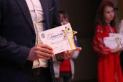 На передньому плані зображено чоловіка в темно-синьому костюмі, білій сорочці та краватці. Він тримає в руках грамоту та статуетку у формі зірки. На грамоті видно герб міста Вараш та напис "Гордість Вараської громади". Також видно підпис та дату "Вараш, 2025".   На задньому плані видно двох дітей. Дівчинка справа одягнена в червону вишиту сукню та вінок на голові, вона також тримає в руках подібну оформлену грамоту. Інша дитина стоїть трохи позаду та лівіше, її видно частково.