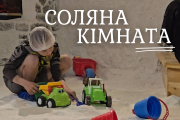 На фотографії зображено інтер'єр соляної кімнати. Стіни та підлога кімнати вкриті шаром солі, що створює відчуття перебування на пляжі з білим піском або в печері. У лівій частині кадру на колінах сидить дитина у шапочці для волосся. Він грається різнокольоровими іграшковими машинками та совочком на соляній підлозі. Поруч з ним розкидані інші дитячі іграшки, зокрема червоне відерце, червона лійка та ще одна машинка.  У верхній центральній частині зображення великими білими літерами написано українською мовою: "СОЛЯНА КІМНАТА".