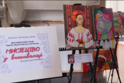 На фотографії зображено виставку дитячих художніх робіт під назвою "Мистецтво у вишиванці". Виставка проходить у Вараській міській раді. На передньому плані праворуч розташовані три картини, встановлені на мольбертах. Перша картина (ближче до глядача) зображує портрет дівчини у вишиванці з червоними квітами у волоссі. Вона тримає в руках білого птаха. Тло картини світле. Друга картина (за першою) виконана в більш абстрактному стилі. На ній зображена фігура в одязі, що нагадує вишиванку, з елементами орнаменту. Кольорова гама картини насичена, з використанням рожевих, зелених, блакитних та жовтих кольорів. Третя картина (на задньому плані) також є портретом дівчини в головному уборі, що нагадує хустку або очіпок, та одязі з елементами вишивки. Кольори на цій картині більш приглушені, переважають рожеві та фіолетові відтінки. На задньому плані видно частину приміщення, схожого на коридор або виставкову залу з бежевими стінами та дверима в кінці. Освітлення в приміщенні природне, з вікон праворуч падає світло.