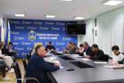 На фотографії зображено засідання виконавчого комітету Вараської міської ради. У просторій кімнаті за довгим столом, вкритим темно-сірою поверхнею, сидять люди. На чолі столу, ближче до центру кадру, сидить міський голова Вараша Олександр Мензул. Він уважно дивиться на когось за кадром.  По обидва боки від нього та вздовж столу розмістилися інші члени виконавчого комітету. Серед присутніх як чоловіки, так і жінки різного віку, одягнені в повсякденний або діловий одяг. Вони зосереджено слухають, роблять нотатки або переглядають документи, що лежать перед ними на столі.  На задньому плані видно стіну, оформлену синім банером з логотипом та написом "Вараська міська рада Varash city council", що повторюється. Також на стіні розміщено герб України, прапор Вараша та, можливо, інші офіційні символи. Праворуч на стіні висить плаский телевізор. Освітлення в кімнаті яскраве, штучне, від стельових світильників.
