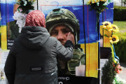 Жінка в хустці та темному пальті ніжно торкається обличчя на портреті загиблого військового, вшановуючи пам’ять сина біля стенду з написом «Героям Слава!» та державним прапором України, прикрашеного квітами та лампадками