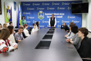 На фотографії зображено зустріч у залі засідань Вараської міської ради. У центрі кадру, за довгим столом, стоїть чоловік у темному костюмі та світлій сорочці - міський голова м.Вараш Олександр Мензул. За столом сидять молоді люди - призери та переможець конкурсів, а також їхні керівники.  На задньому плані видно синій банер з написом "Вараська міська рада" та гербом міста, а також державний прапор України та прапор міста Вараш. На стіні також розміщено великий телевізор. Загальна атмосфера зустрічі є позитивною та святковою.