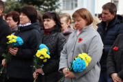 Кілька людей різного віку стоять, тримаючи букети з жовтими та блакитними квітами. На одязі деяких присутні червоні маки. Вирази їхніх облич зосереджені та дещо сумні.